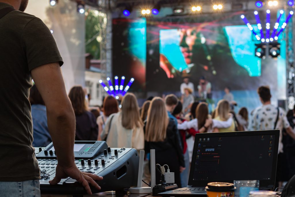 Technicus bedient apparatuur voor professionele beeld- en geluidsoplossingen tijdens een live evenement, met publiek en podium op de achtergrond.