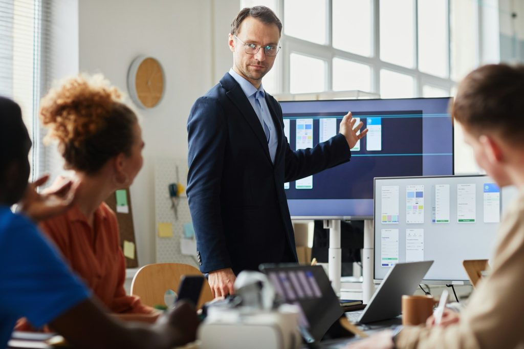 Zakelijke presentatie met man in pak die gegevens toont op een scherm aan een team tijdens een vergadering, ondersteund door professionele audiovisuele oplossingen met laptops en visuele hulpmiddelen.
