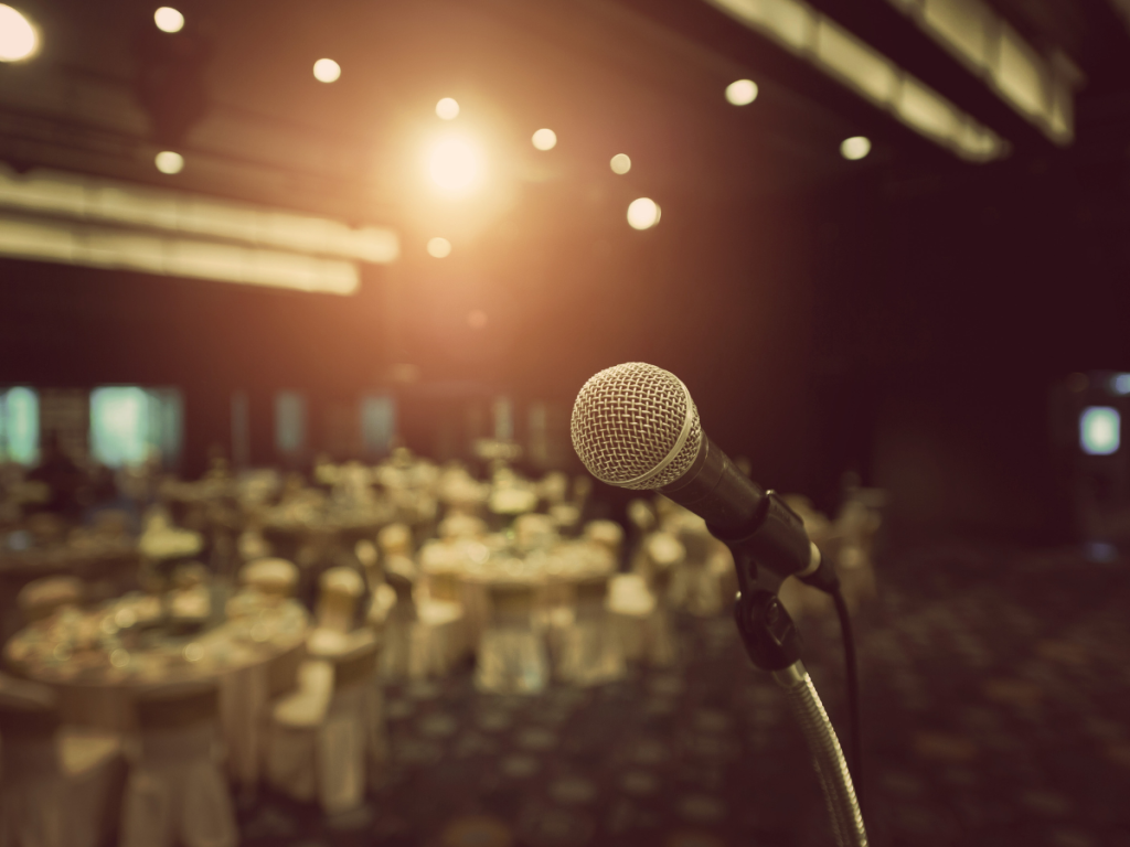 Een spreker presenteert op het podium tijdens een audiovisuele productie evenement, met krachtige verlichting en een betrokken publiek op de achtergrond.