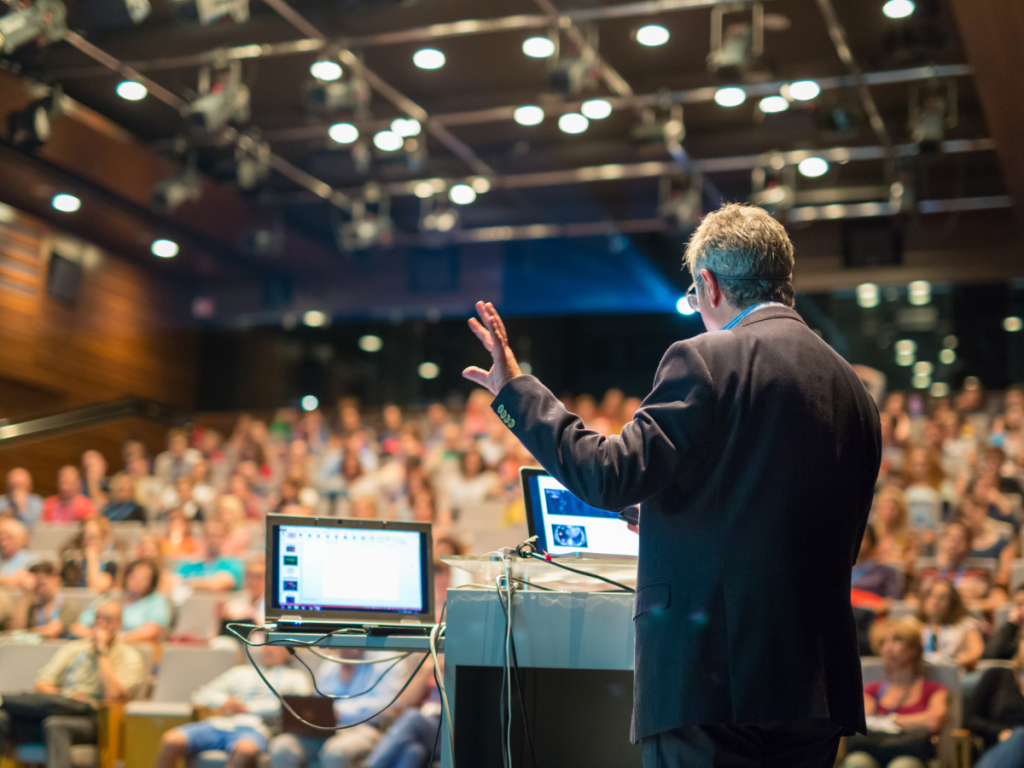 Een spreker presenteert tijdens een zakelijk evenement, ondersteund door een professioneel geluidssysteem voor evenementen in een goed verlichte conferentiezaal.