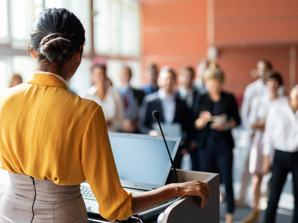 Een vrouw in een gele blouse spreekt voor een professioneel publiek tijdens een zakelijke bijeenkomst. Op de voorgrond staat een microfoon en een laptop, wat duidt op een formele presentatie. De luisteraars zijn netjes gekleed en gefocust, wat wijst op een conferentie of seminar. Professionele audiovisuele ondersteuning met helder geluid en een goed zichtbaar scherm is essentieel om een effectieve presentatie te garanderen.