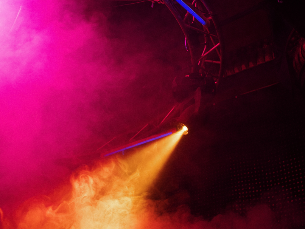 Kleurrijke podiumverlichting met een spotlight die een warme oranje gloed uitstraalt, omringd door roze en paarse lichteffecten en rook. De dynamische sfeer benadrukt een professionele lichtshow voor evenementen of concerten.