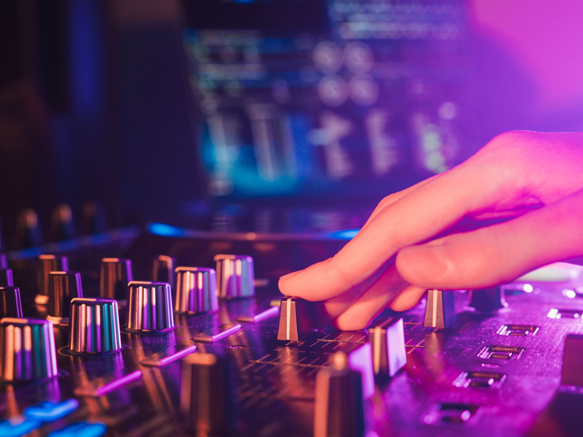 Close-up van een DJ-mixer met kleurrijke LED-verlichting en een hand die de knoppen aanpast. De professionele geluidsapparatuur zorgt voor een kristalheldere audio-ervaring, essentieel voor live muziek, evenementen en optredens. De combinatie van geluid en verlichting creëert de ultieme sfeer voor een energiek feest of een spectaculaire show.